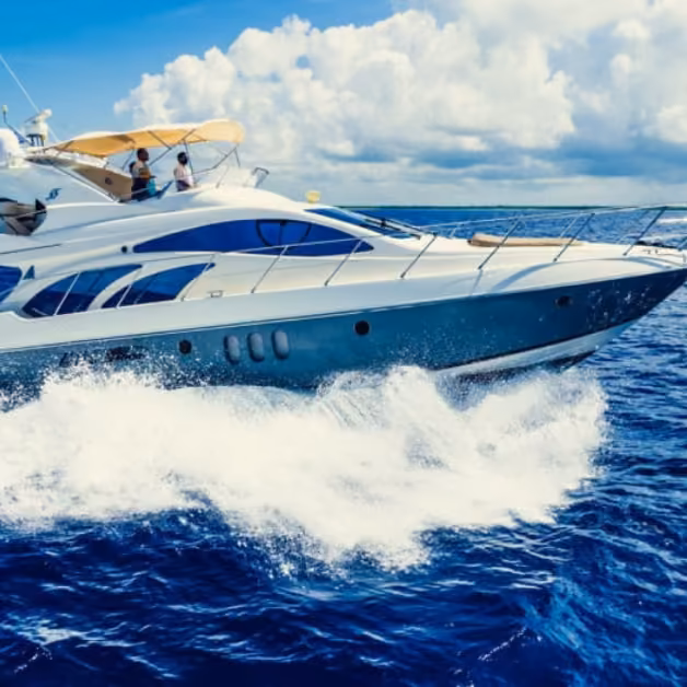 A white yacht glides across vivid blue water, leaving waves as two people relax on deck under a partly cloudy sky.