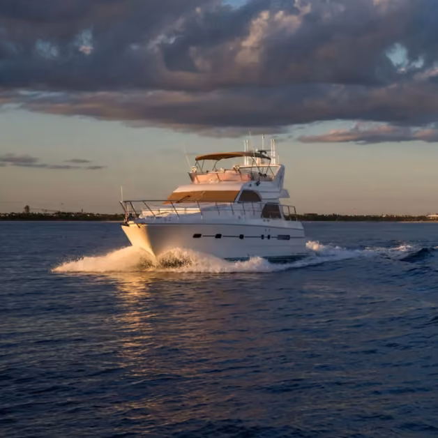 A white yacht cruises on calm blue water beneath a cloudy sky, sunlight casting a warm glow across the vessel.