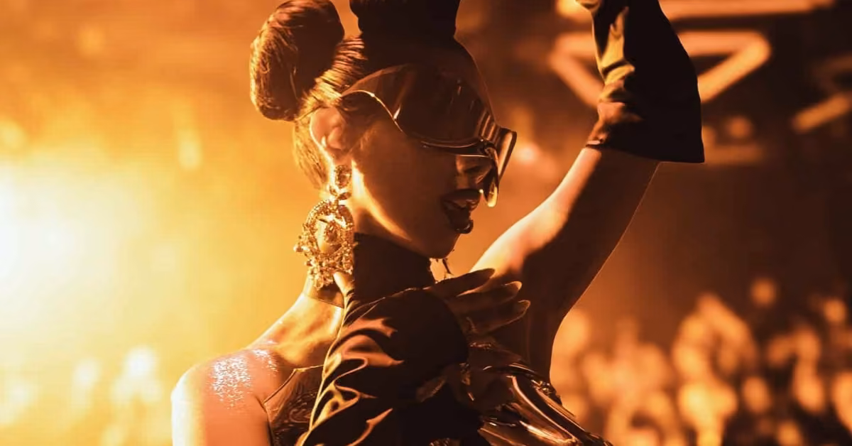 A glamorous performer in futuristic attire, large earrings, and sunglasses poses onstage under golden lights before a blurred audience.