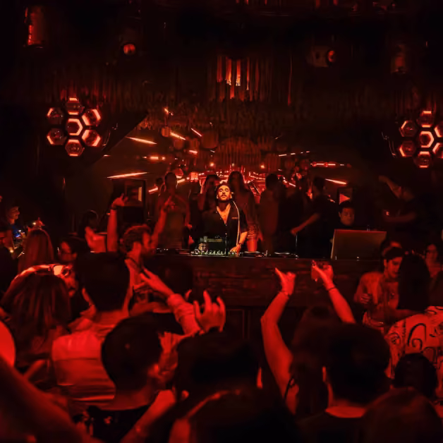 A DJ plays on stage in a crowded night club lit by red and orange lights, while people dance in the lively atmosphere.