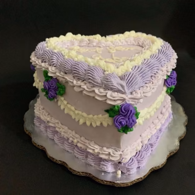 Heart-shaped cake with purple and white frosting, piped borders, small rose-like flowers on a silver board against black.