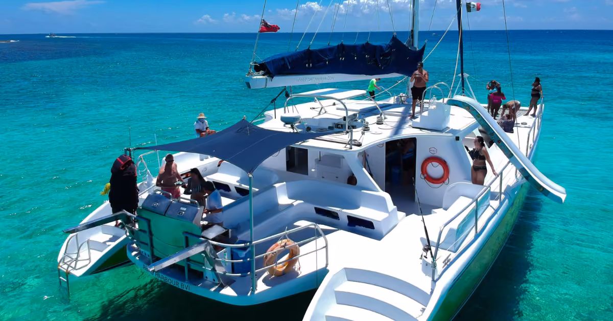 People relax and enjoy the sun on a white catamaran with a slide, floating on clear turquoise water under a bright blue sky.