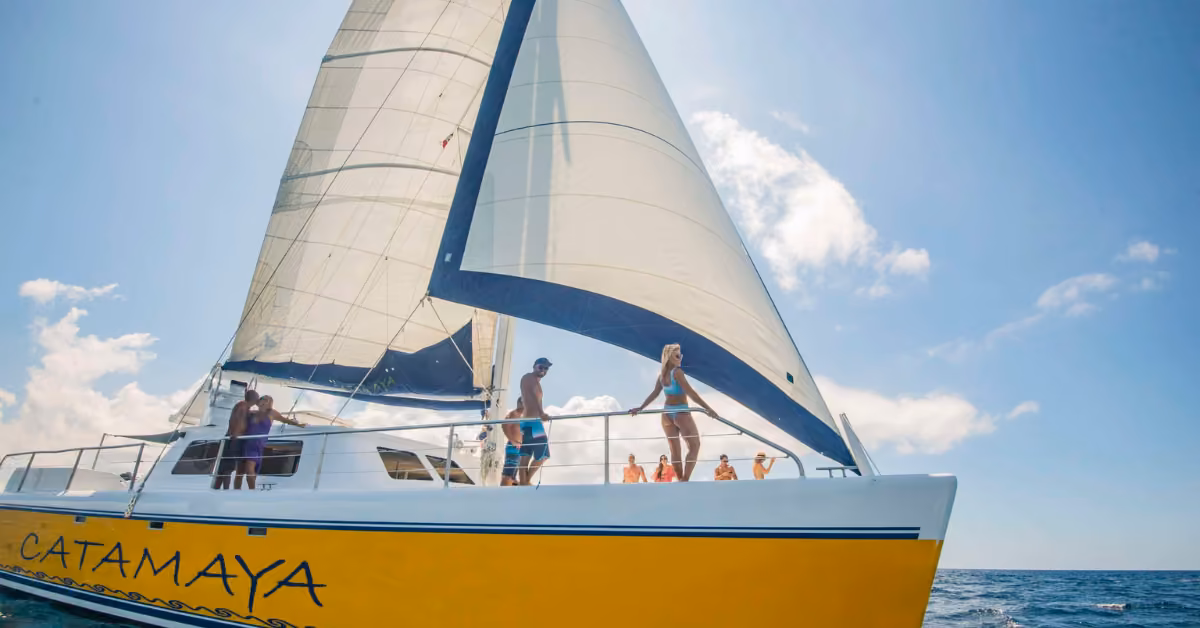 A yellow and white sailboat named Catamaya glides on the ocean as guests enjoy views from the deck under a partly cloudy sky.