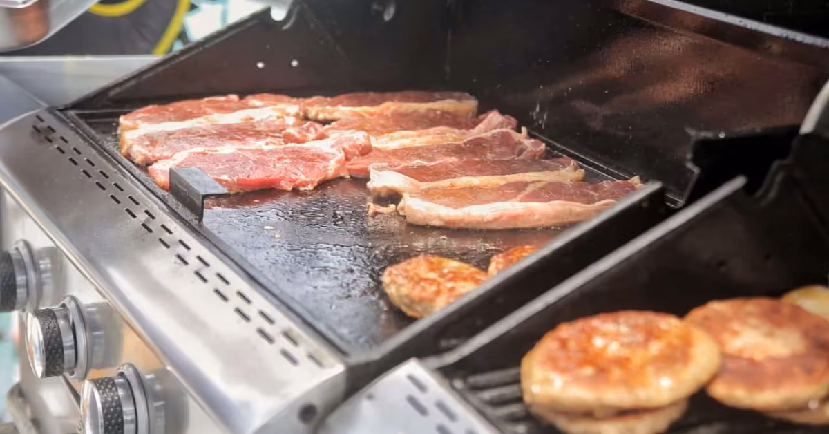Steaks and burger patties cooking on a gas grill, with patties to the right and steaks spread across the flat top surface.