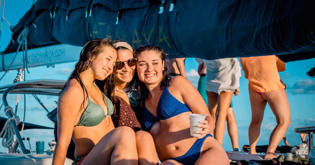 Three young women in swimsuits smile together on a catamaran under sunny blue skies. One holds a white cup; others are nearby.