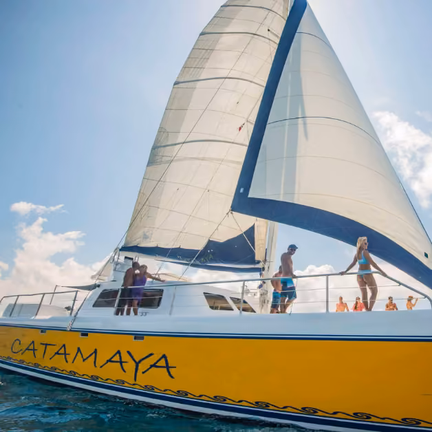 The yellow Catamaya catamaran sails on a sunny day as people in swimwear relax on deck under its white sails.
