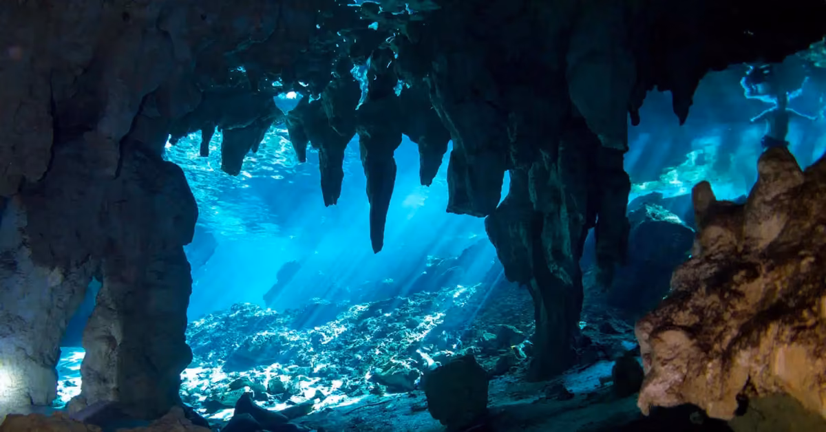 Sunlight filters through water, lighting up stalactites and rocks inside a blue-green underwater cave.