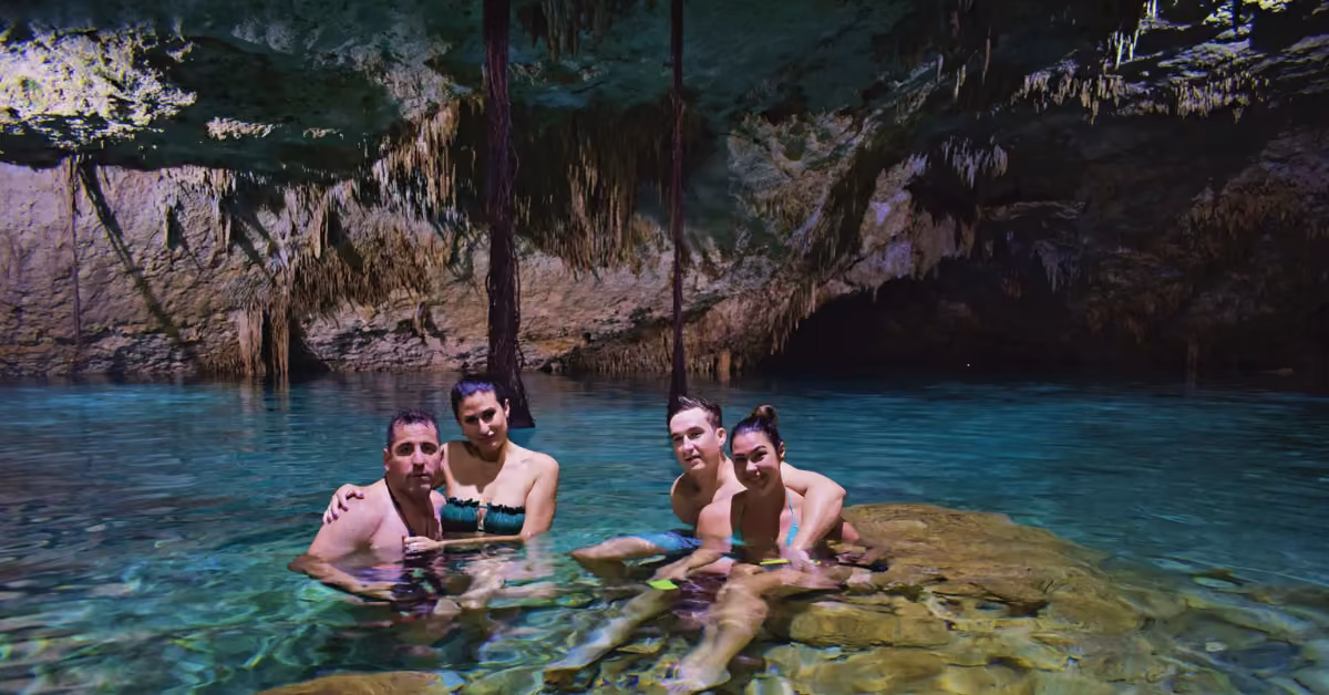 Four people in swimsuits relax in clear water inside an underground cave, surrounded by rock walls and hanging stalactites.