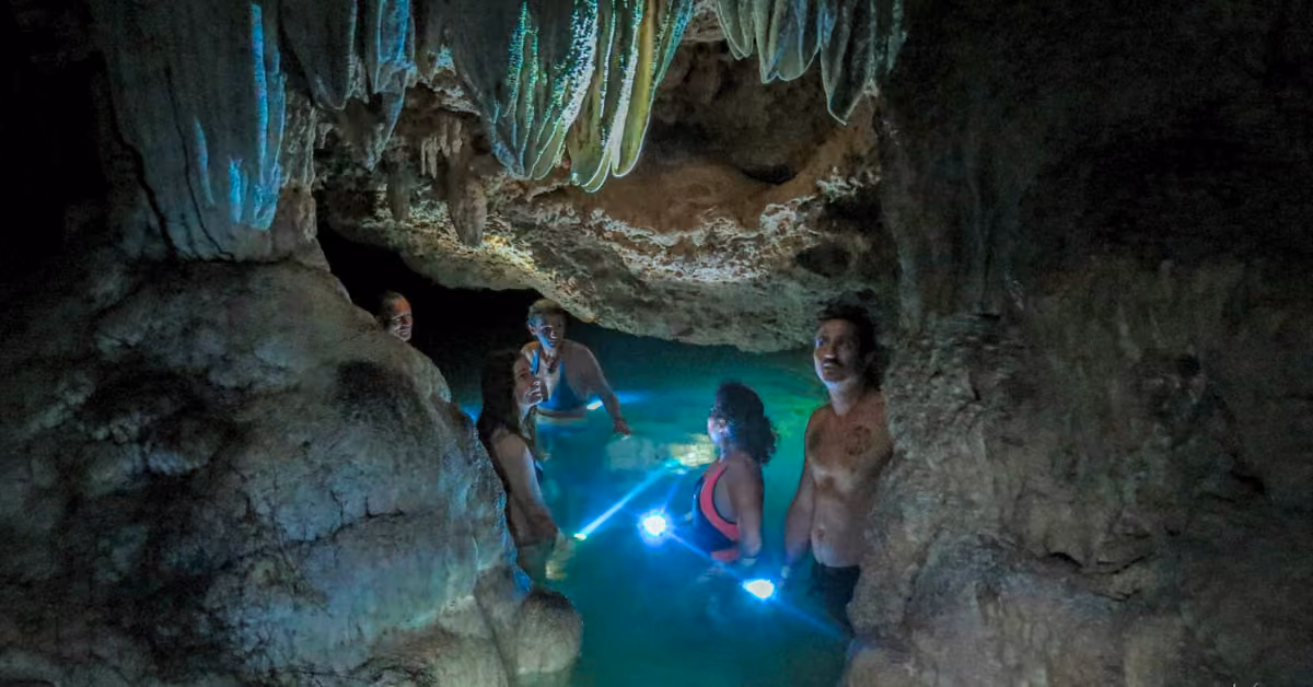 Four people wade through turquoise water in a dark cave, flashlights shining as stalactites hang above.