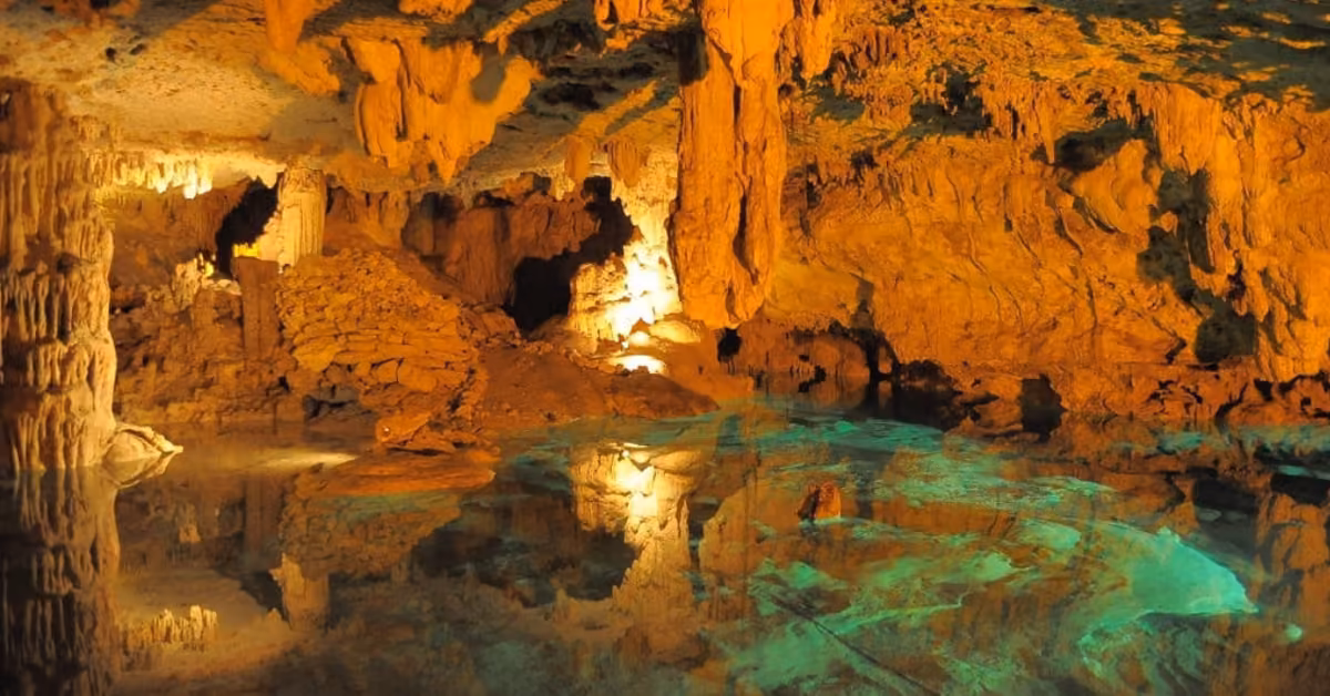 Sunlit cave with orange-brown stalactites and stalagmites mirrored in a turquoise pool, creating a peaceful, mystical scene.