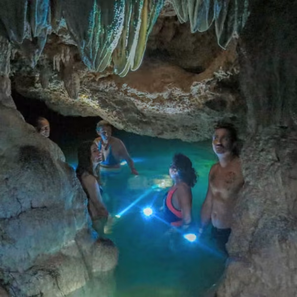Five people waist-deep in turquoise cave water, two holding lights that brightly illuminate rocky walls and hanging formations.
