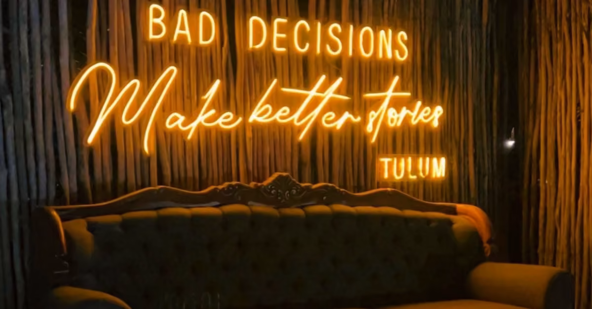 A neon sign at Chambao Restaurant reads BAD DECISIONS Make better stories TULUM above a green vintage sofa against a dark wall.