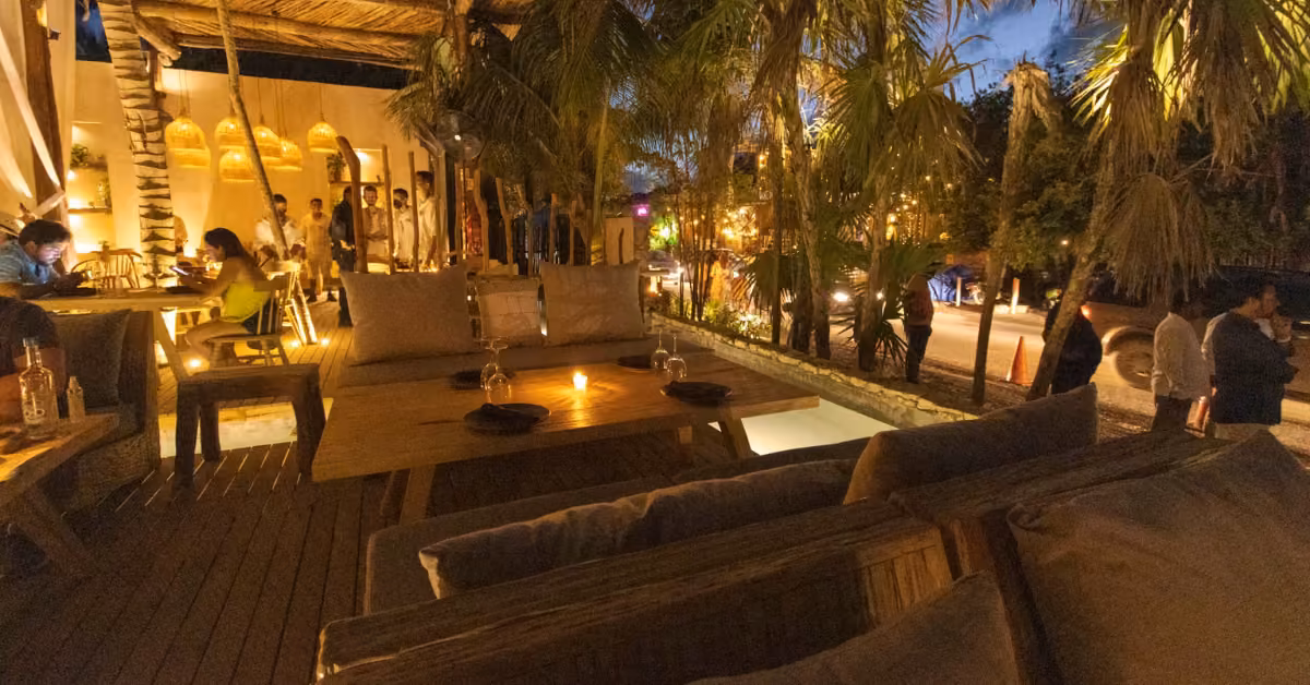 Chambao’s patio at dusk with cushioned seats, wooden tables, warm lights, palm trees, and diners socializing by the street.