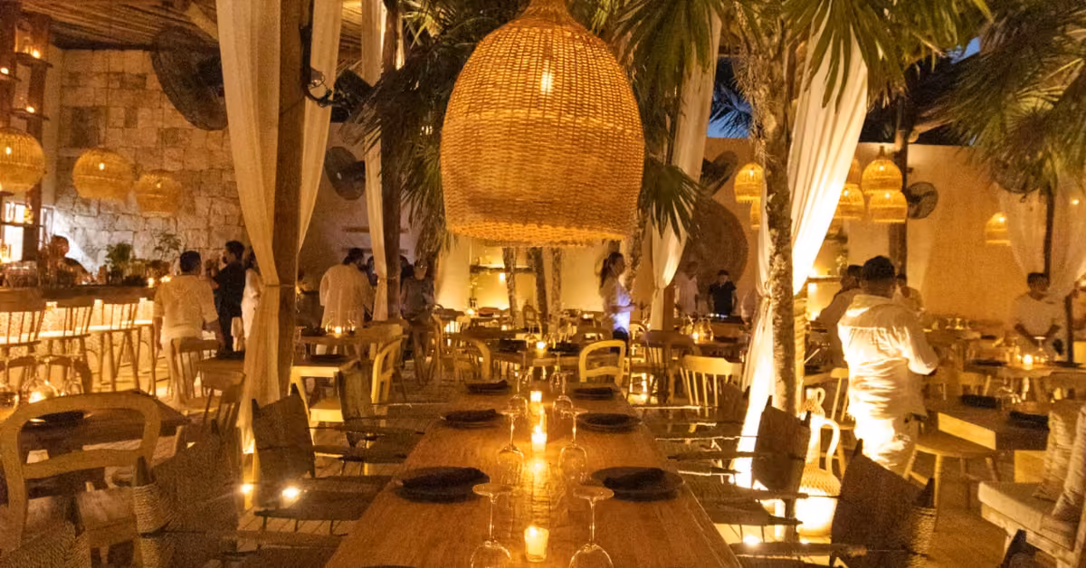 A warmly lit restaurant with wicker lamps, palm trees, and white curtains; diners enjoy a relaxed, tropical evening setting.