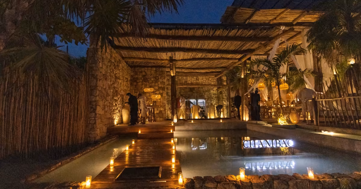 At night, people mingle at a cozy outdoor restaurant with stone walls, wooden beams, and candlelit poolside ambiance.