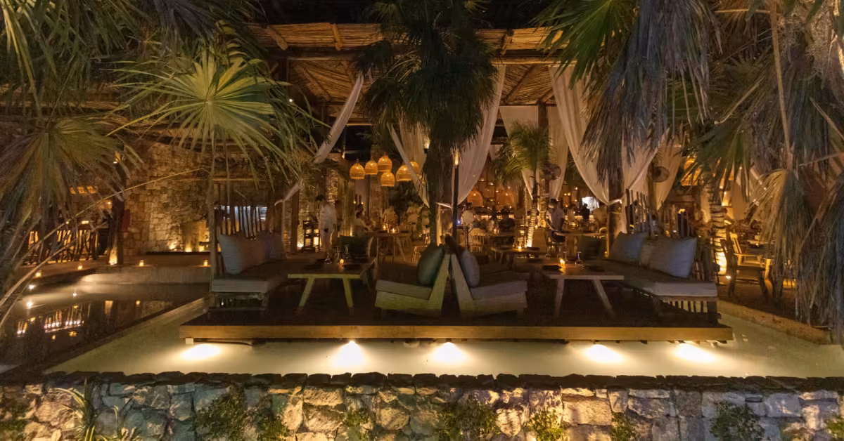 Chambao Restaurant’s outdoor lounge features cozy seats, soft lighting, white curtains, palm trees, and a shallow pool at night.