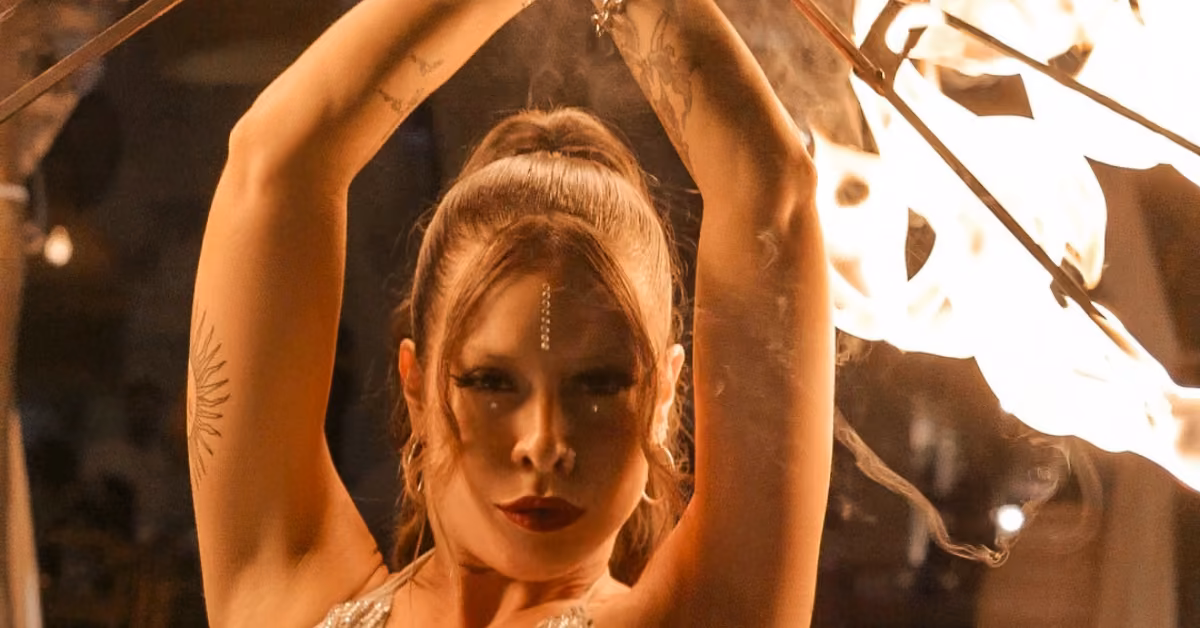 A tattooed woman performs a fire dance, arms raised with flaming sticks, her sparkly makeup glowing in the firelight.
