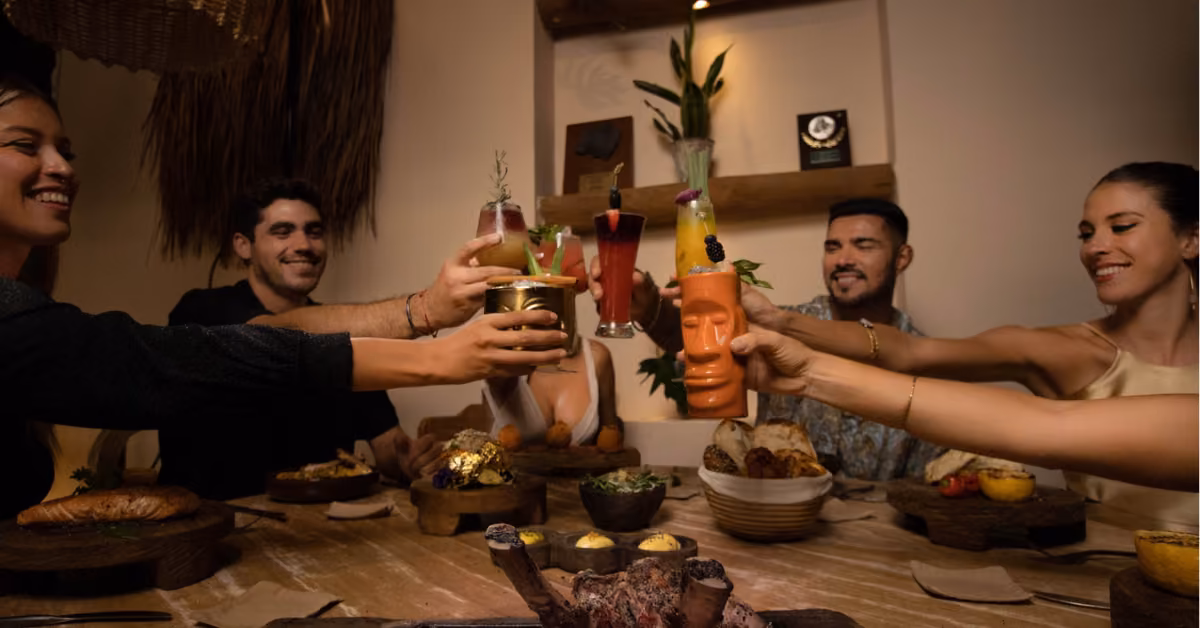 Four friends toast with colorful drinks, smiling around a table of food in the warm, cozy glow of Chambao restaurant.