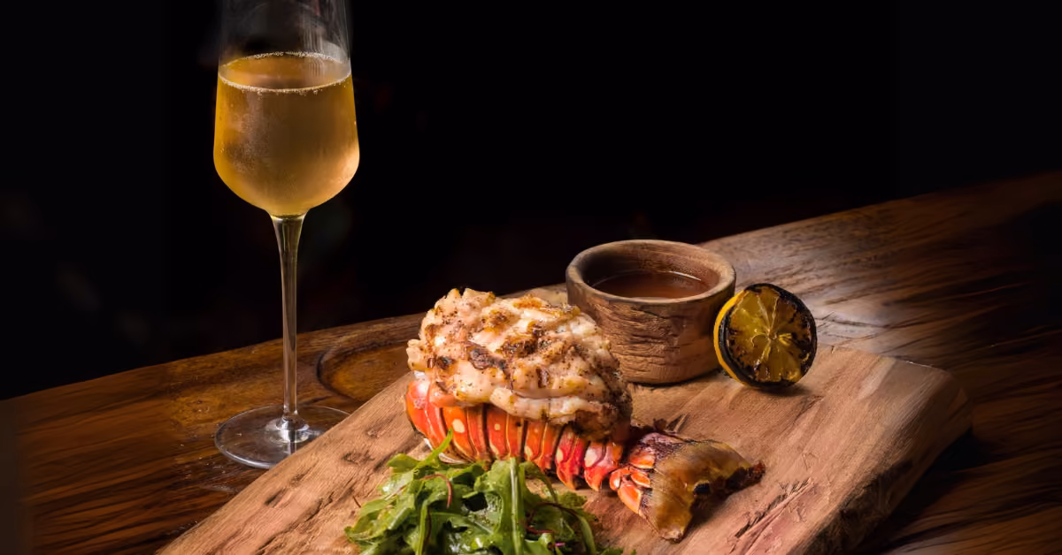 Grilled lobster tail on a wooden board with fresh greens, grilled lemon, sauce, and white wine at a rustic restaurant table.
