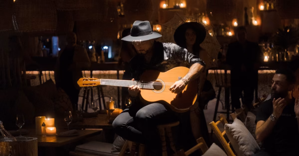 A musician in a black hat and patterned shirt plays acoustic guitar in the cozy, candlelit Chambao restaurant as guests watch.