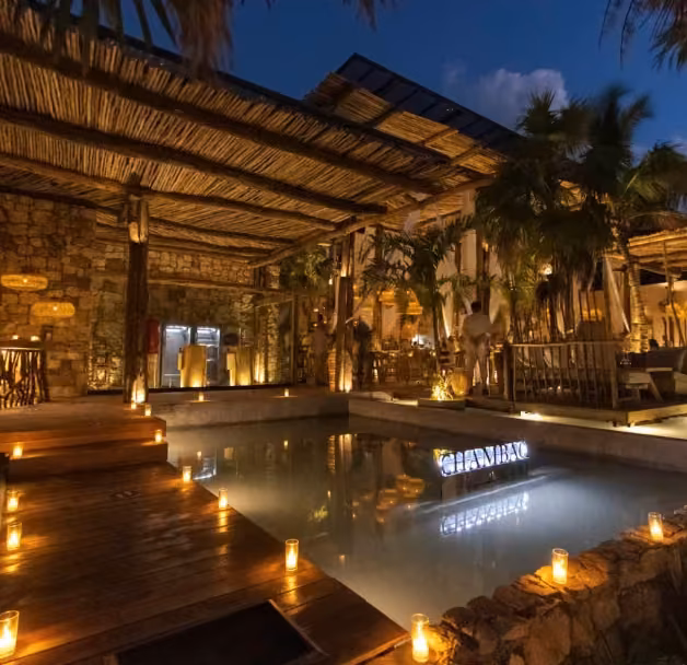 Outdoor restaurant at night with a stone building, palm trees, and candlelight by a pool, creating a cozy, inviting scene.
