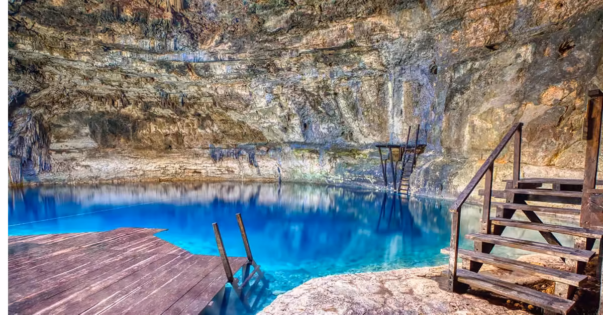 Sunlight streams into a cenote cave with turquoise water, jagged stalactites, and a wooden platform over the shimmering pool.