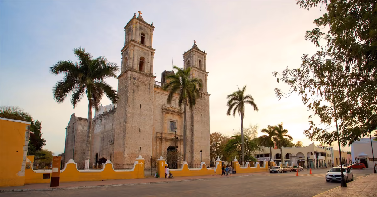 Colonial stone church with twin towers behind a yellow wall, palm trees around, people and cars nearby at sunset.