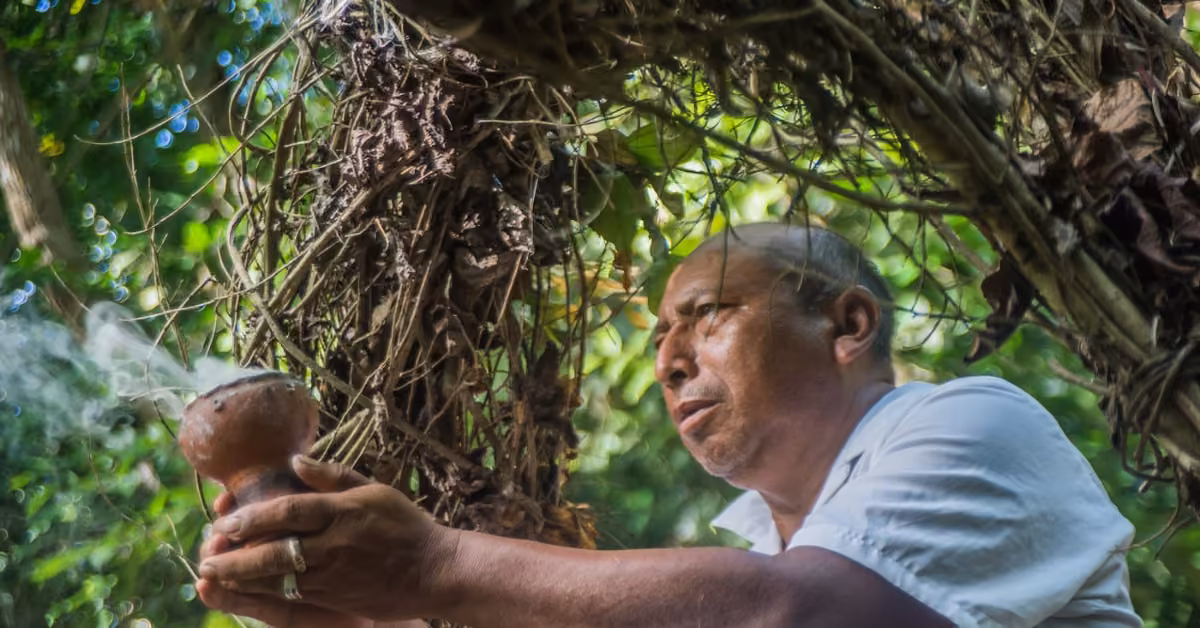 A man in a white shirt holds a smoking clay vessel in lush jungle, sunlight streaming through leaves above him at Coba ruins.