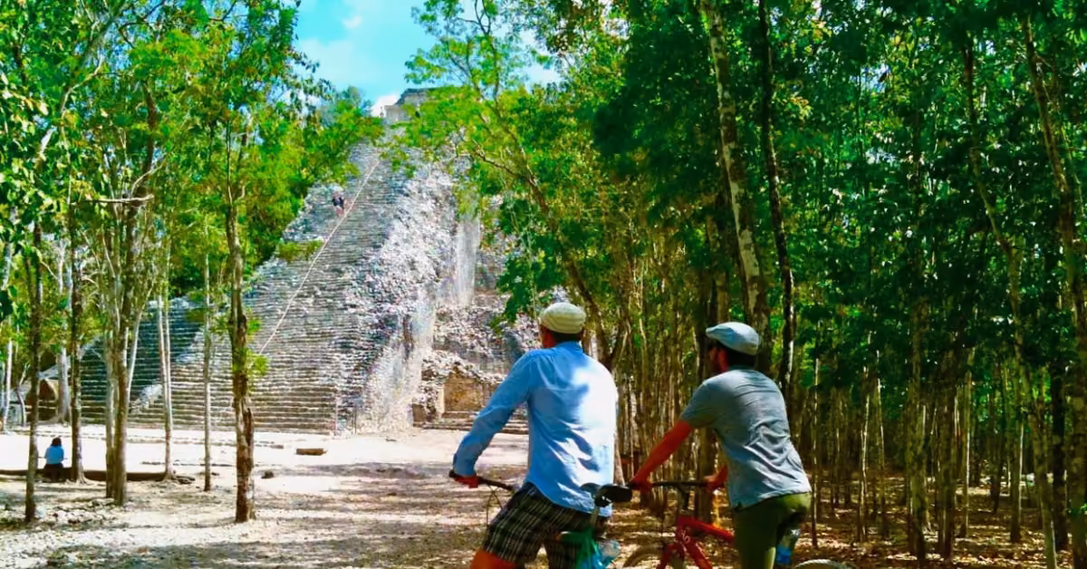 Two cyclists pause to admire an ancient stone pyramid among lush trees under a sunny blue sky at the Coba ruins.