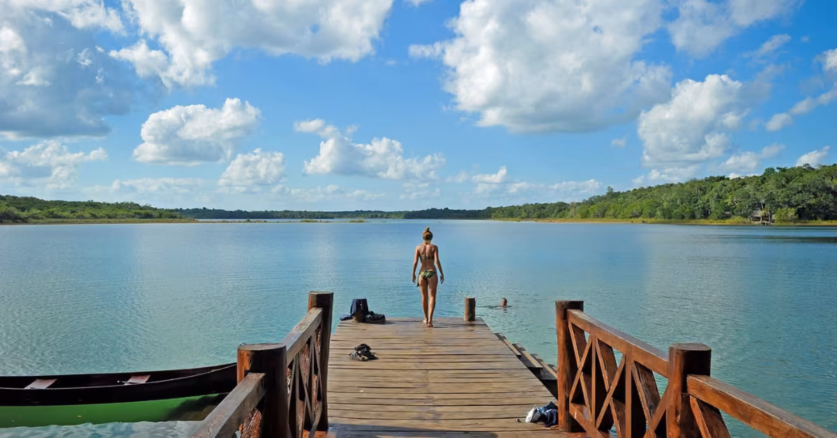 A person in swimwear stands on a wooden dock overlooking a calm lake, with greenery, clouds, a canoe, and another swimmer nearby.