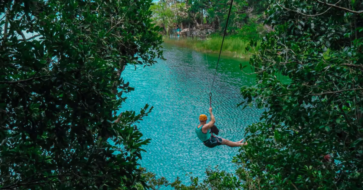 A person in a helmet ziplines over a turquoise river, surrounded by lush green trees, enjoying an exciting outdoor adventure.