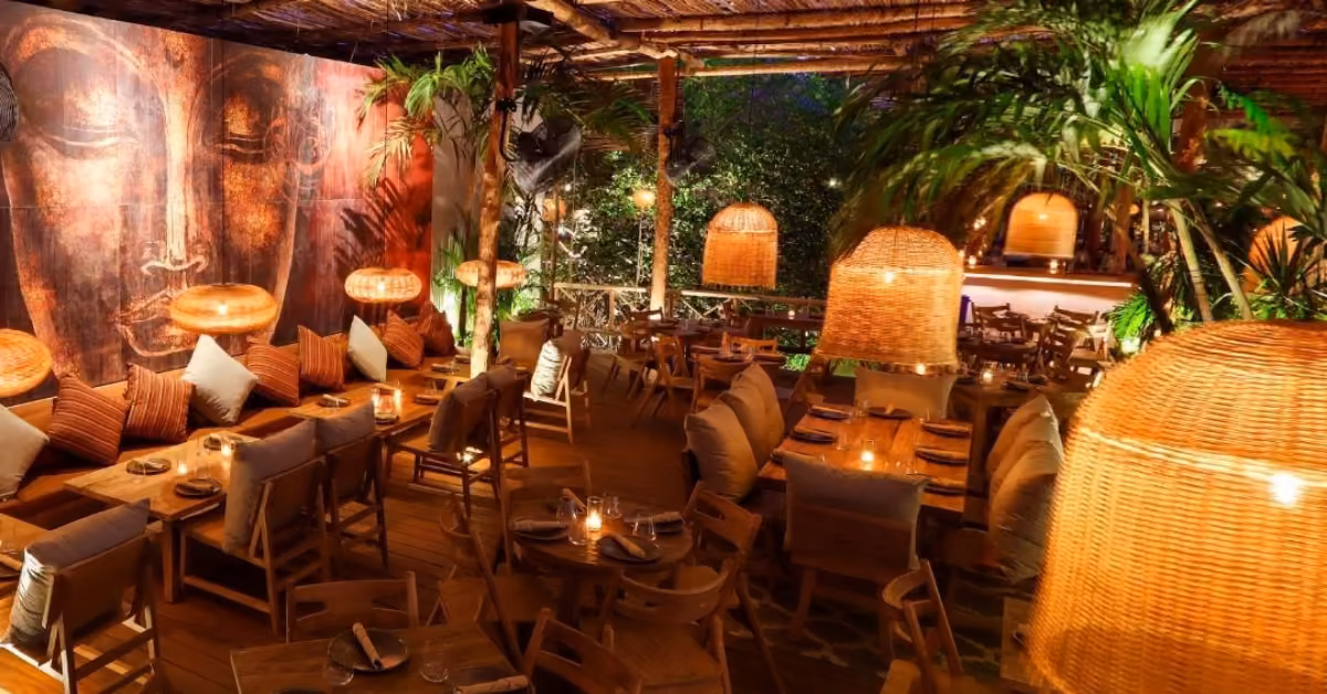Warmly lit restaurant with wooden tables, rattan lamps, lush plants, and a serene face mural, creating a cozy vibe.