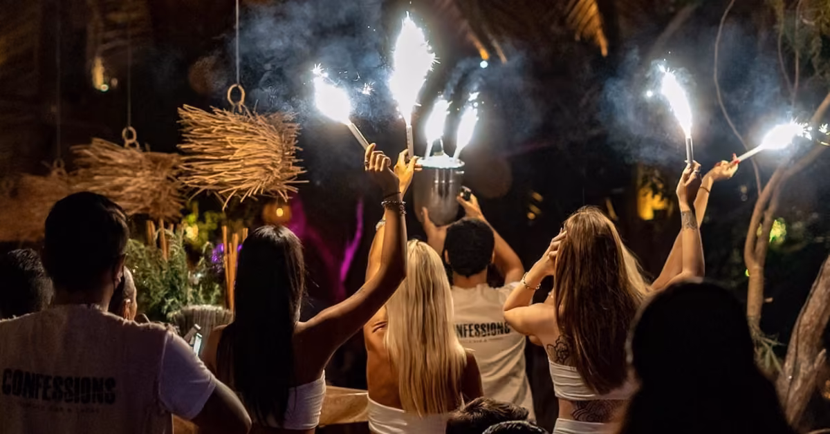 People in a festive, dim outdoor setting hold sparklers amid lush greenery, some wearing matching shirts, celebrating together.