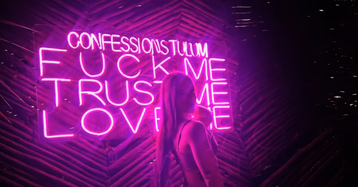 A person stands before a neon pink sign reading “FUCK ME TRUST ME LOVE ME,” bathed in vivid pink light at a night club.