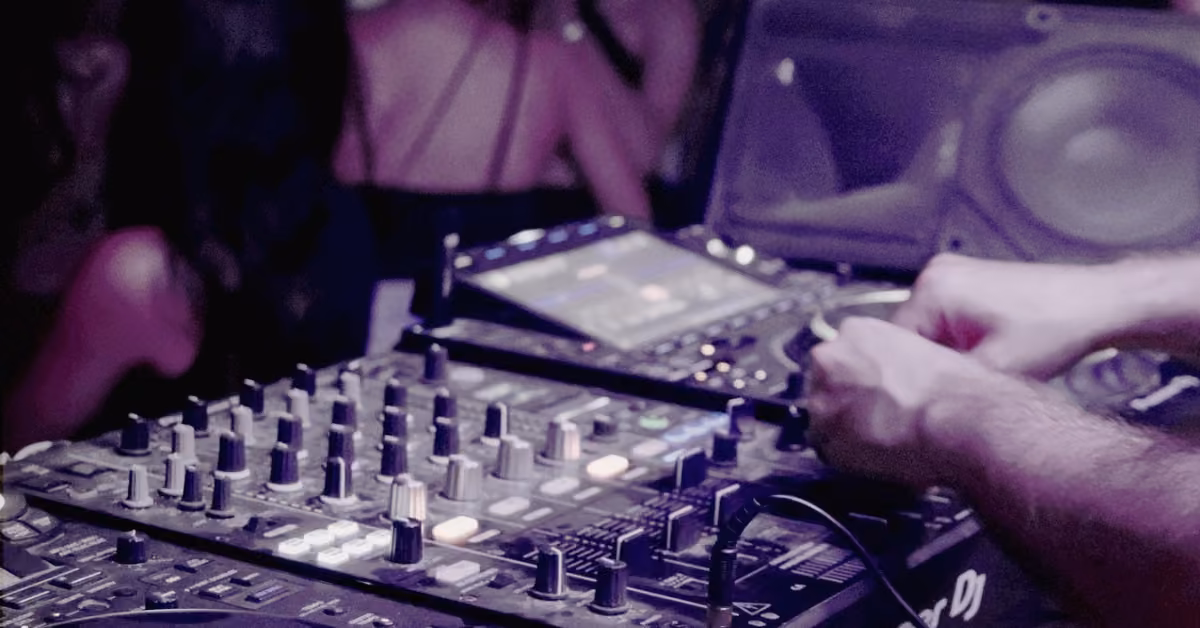 DJ’s hands adjust controls on a mixer and turntable at Confessions Night Club, dancers blurred under colorful lights.