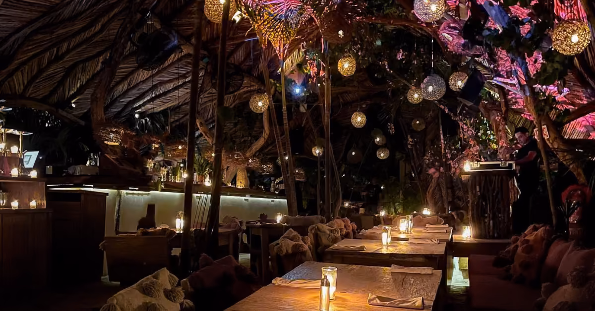 A dim restaurant with wooden tables, cozy seating, lanterns, and lush decor radiates warmth. Candles give a gentle glow.