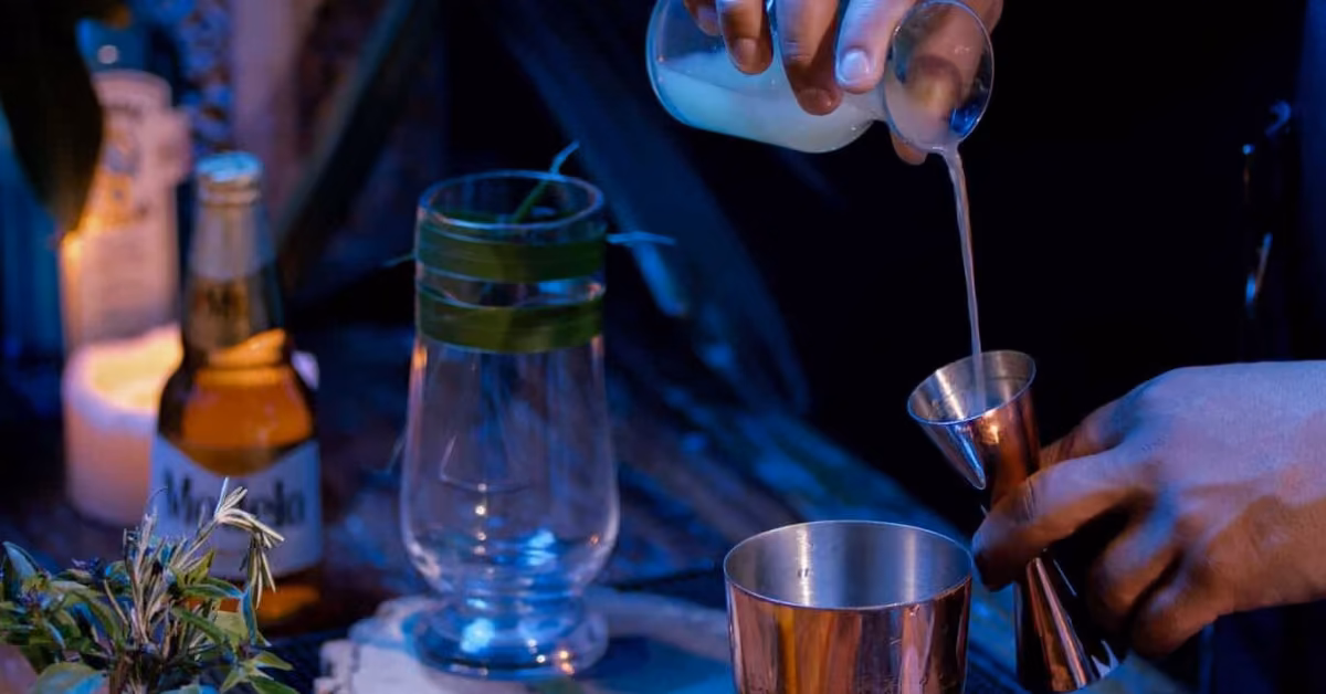 Someone pours a white liquid from a glass jar into a jigger over a shaker. A beer, leafy glass, candle, and plant sit nearby.