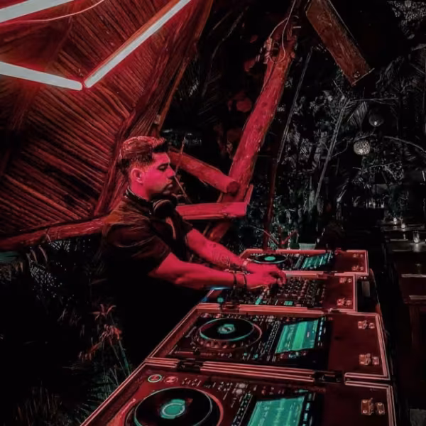 A DJ with headphones mixes music on glowing decks in a dim room lit by neon pink, surrounded by wooden beams and nightlife energy.