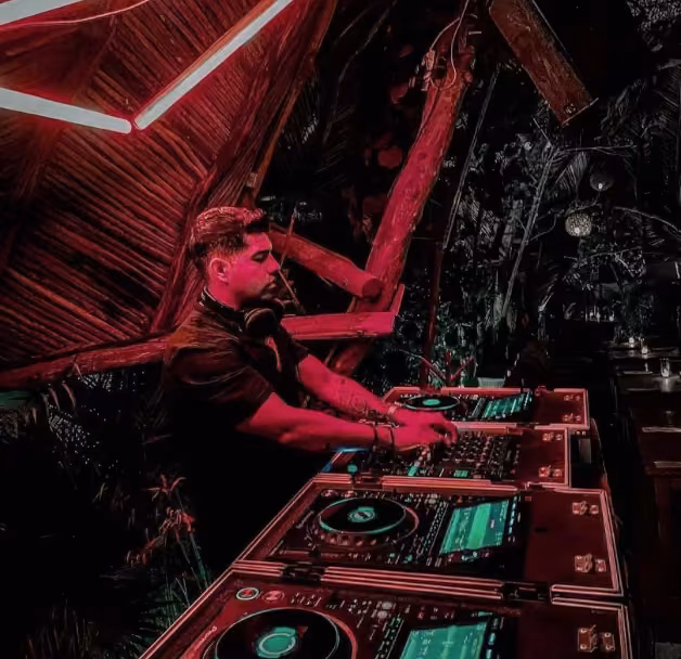 A DJ with headphones mixes music on glowing decks in a dim room lit by neon pink, surrounded by wooden beams and nightlife energy.