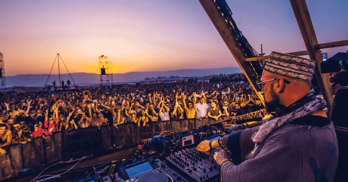A DJ plays on stage at sunset for a lively crowd at Day Zero Festival, with mountains silhouetted in the background.