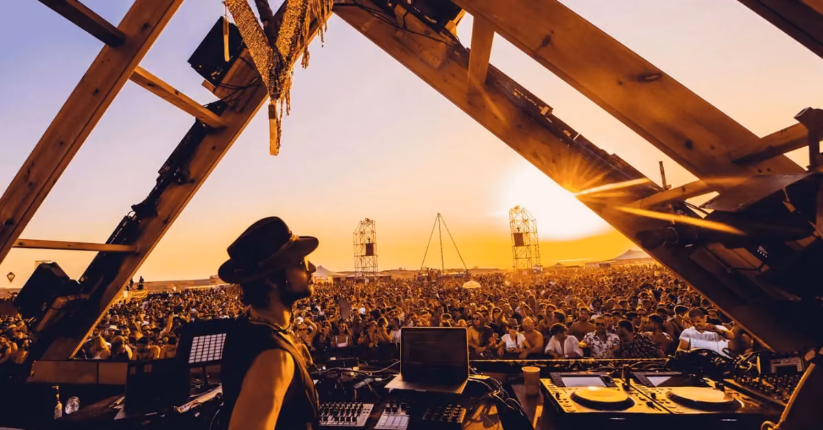 A DJ in a hat plays onstage at sunset at Day Zero Festival, with equipment visible and a crowd enjoying music outdoors.