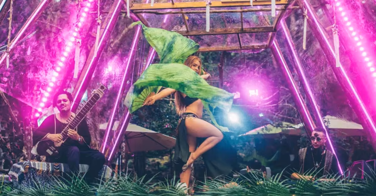 A woman dances with flowing green fabric under purple lights at Day Zero Festival, musicians nearby, lush greenery all around.