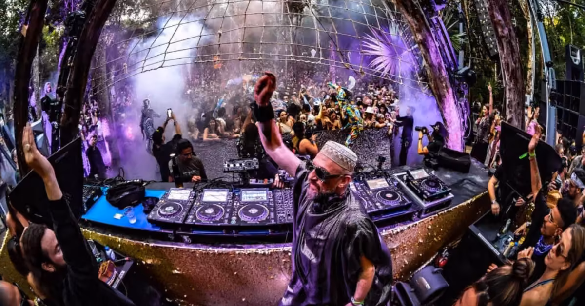 A DJ plays on an outdoor stage at Day Zero Festival as a lively crowd dances under colorful lights and mist among trees.