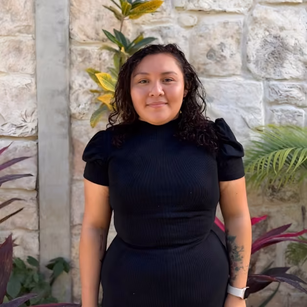 A woman with wavy dark hair in a black dress and white watch stands smiling by a stone wall with green and purple plants.