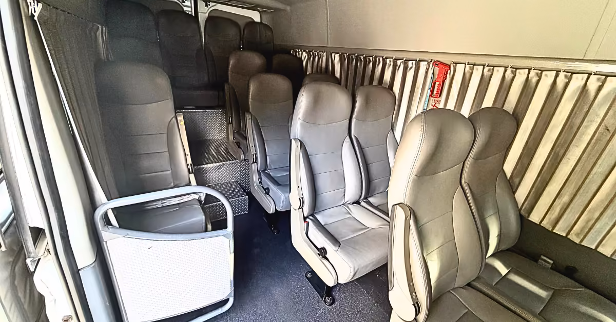 Rows of empty gray seats in a small bus, metal barrier by the door, ribbed walls, and a red fire extinguisher on the wall.