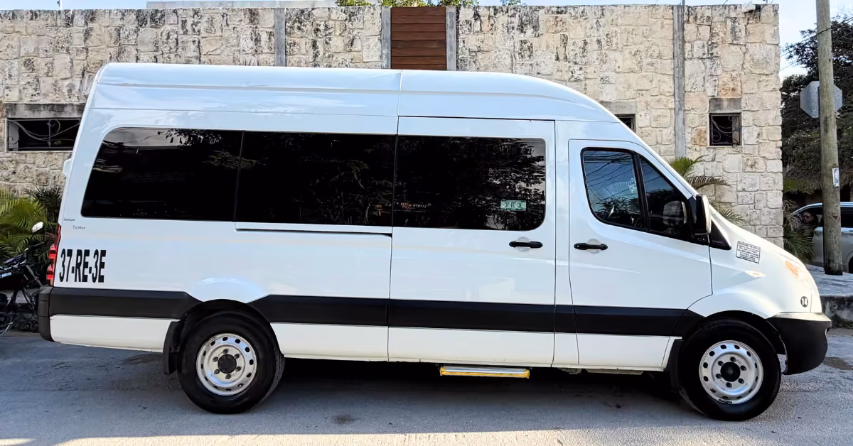 A white van with tinted windows and black trim is parked by a stone wall and greenery. 57/RE3E is marked near the rear.