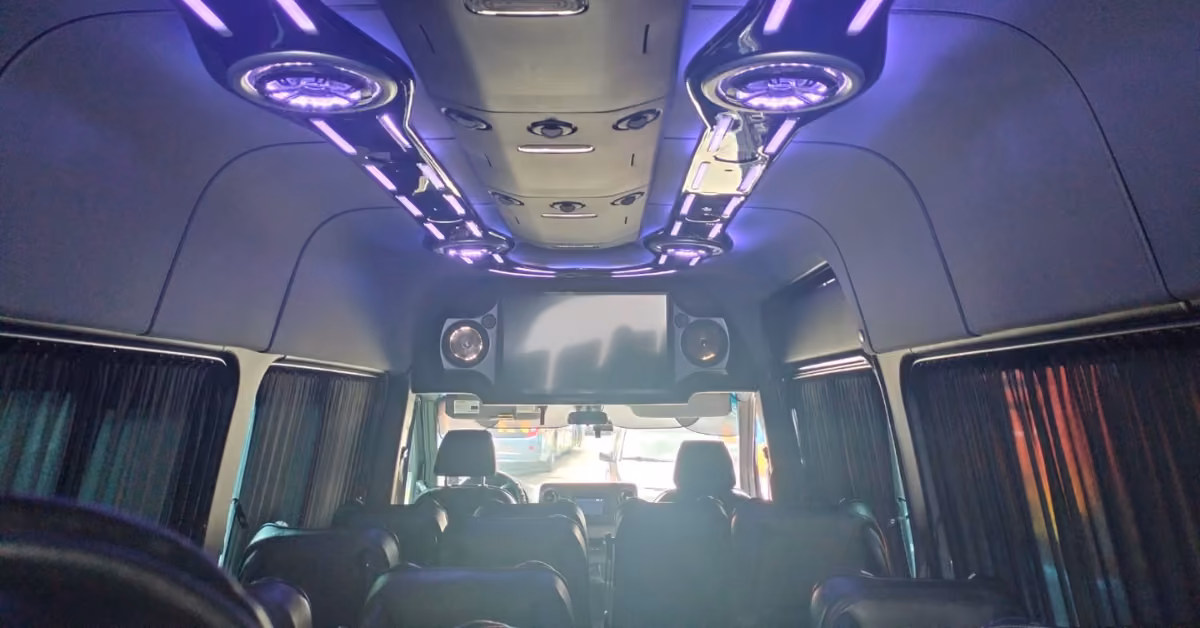 Luxury van interior with black leather seats, tinted curtained windows, purple ceiling lights, speakers, and a front monitor.