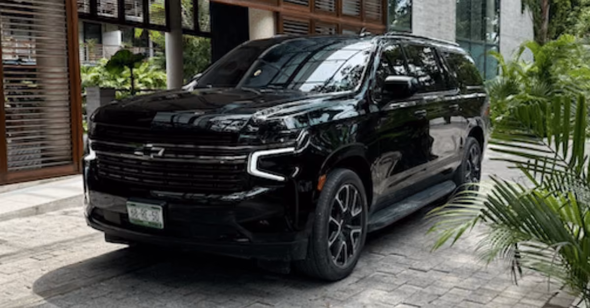 A black Chevrolet Suburban SUV is parked on a stone driveway by a modern building with large windows and green plants.