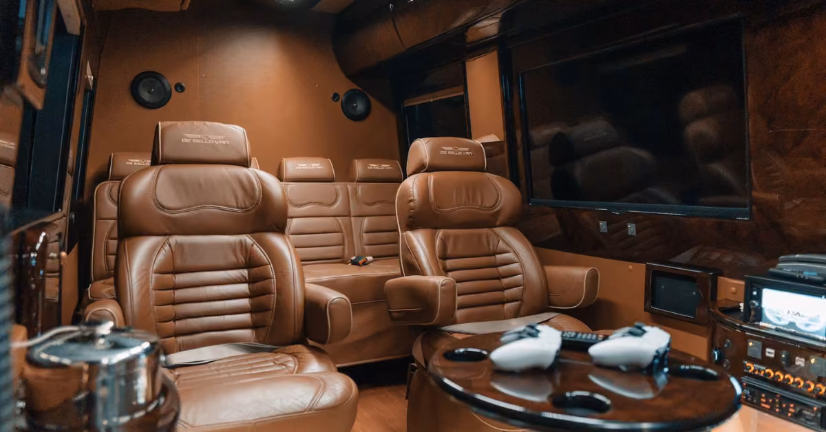 Airport van interior with four brown leather seats, a round table with game controllers, large windows, and built-in screens.