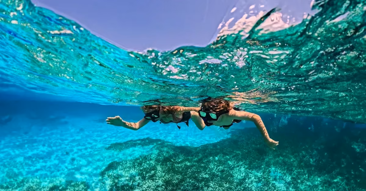 Two people snorkeling in clear turquoise water above a coral reef, sunlight streaming down and blue sky visible above the surface.